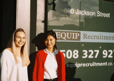 Two young woman standing and smiling in front of a window that says Equip Recruitment